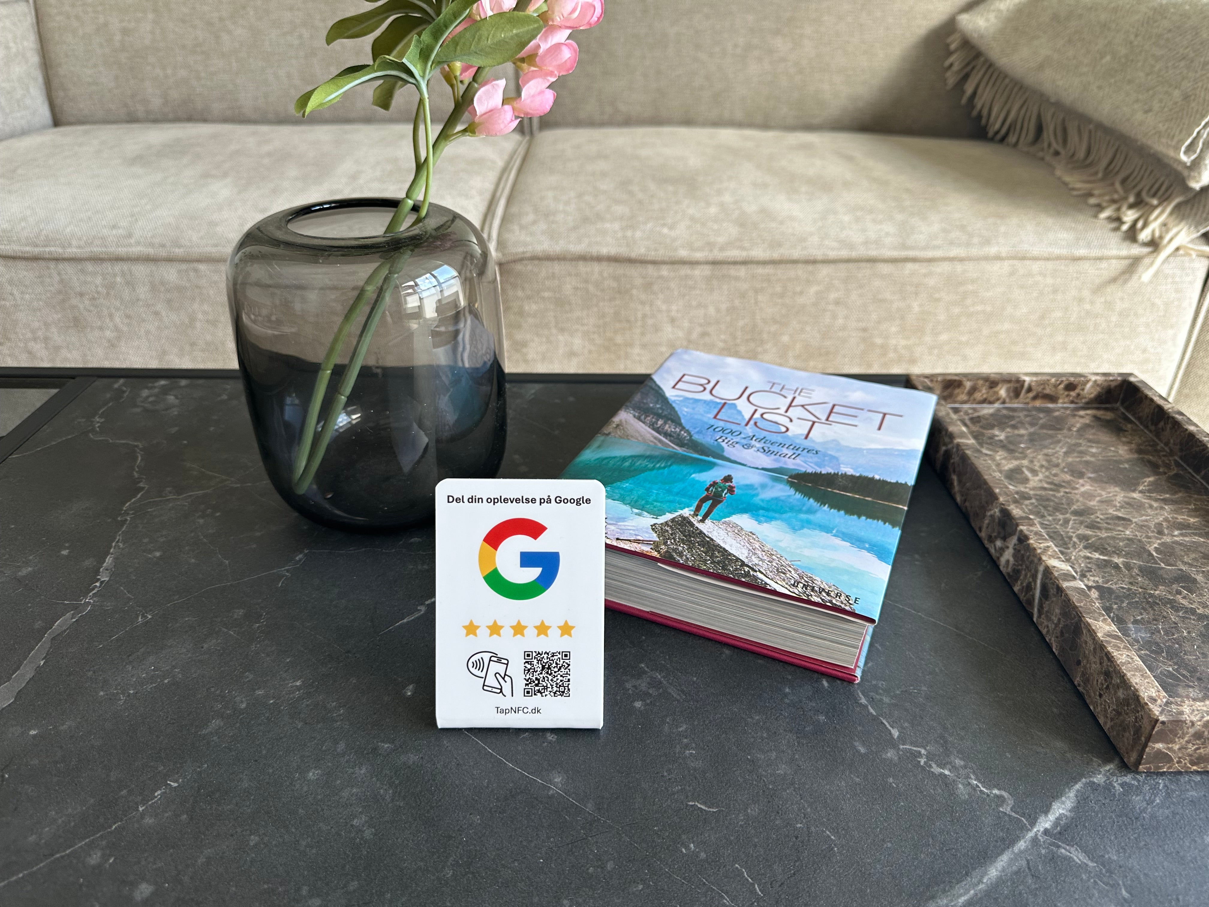 En googleanmeldelses bordstander med NFC og QR kode på et lille bord med en bog og vase.
